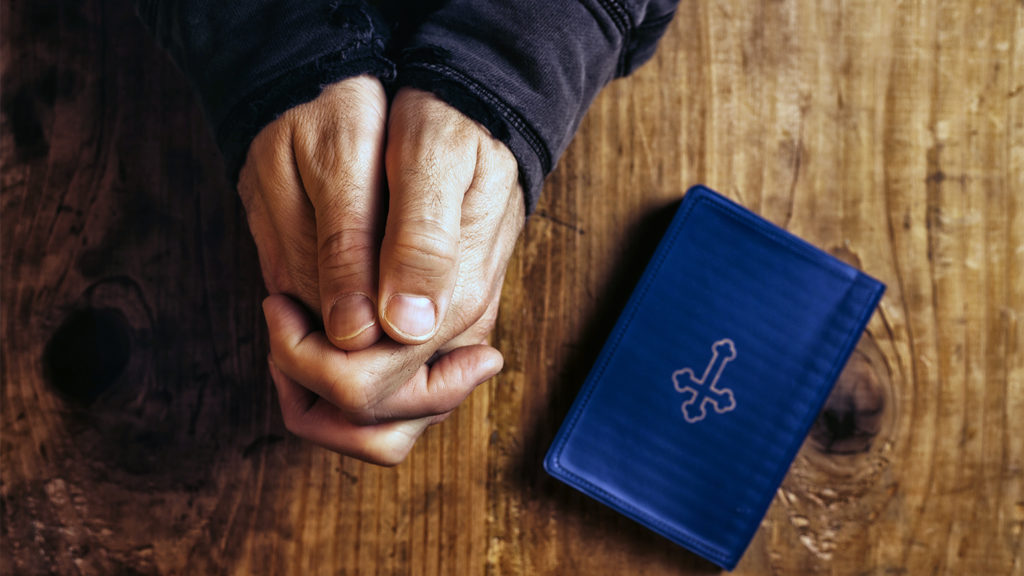 A man's hands clasped in nighttime prayer meditation, with a Bible resting nearby