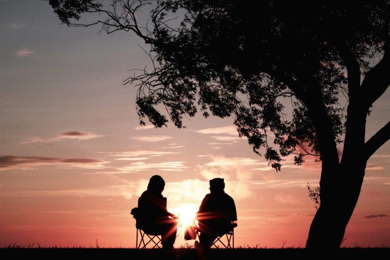 Friends sit outside under a tree at sunset talking about living above offense.